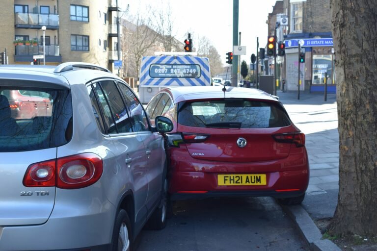 Red hatchback tightly wedged between a parked silver SUV and a sidewalk tree on a city street