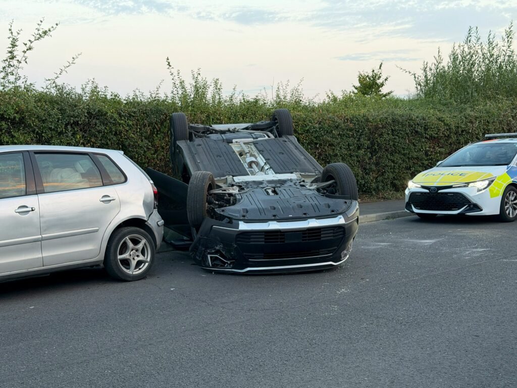 Overturned dark car lying upside down on a road near a silver hatchback and a police vehicle.