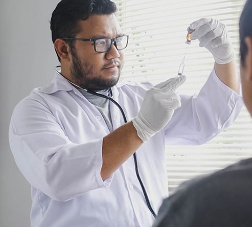 Healthcare professional wearing glasses, a white lab coat, and medical gloves holding up a small glass vial to fill a syringe before administering treatment