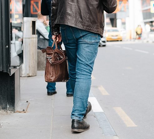 Pedestrian carrying a brown leather bag walking along a city street pavement.