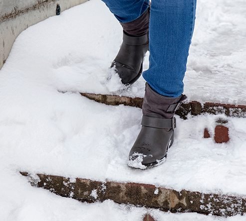 Close-up of a person in blue jeans and dark leather boots descending outdoor stairs covered in fresh snow and ice