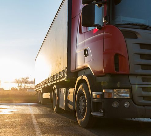 Low-angle view of a red semi-truck with a large cargo trailer traveling on an open road during golden hour