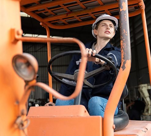 Low-angle shot of a female worker wearing a white hard hat and workwear seated inside the cab of an orange forklift, holding the steering wheel.